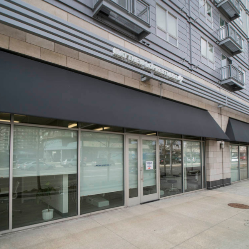 The image shows the exterior of a commercial building with large glass windows and doors, featuring black awnings.