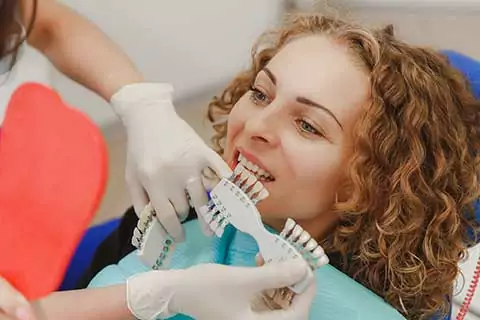Woman in a dental clinic with a shade guide next to their mouth, showing tooth-colored tabs for matching dental restorations.