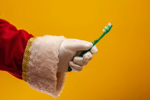 A hand wearing a white glove and a red sleeve with gold trim holds a green toothbrush with white bristles against a solid yellow background. The costume details resemble Santa Claus attire.