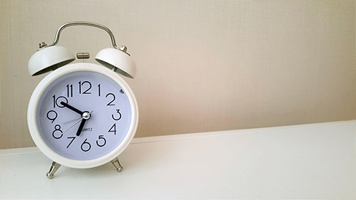 The image shows a white analog alarm clock with two bells on top, placed on a white surface against a beige background. The clock face has black numbers and hands, with the time showing approximately 6:50.
