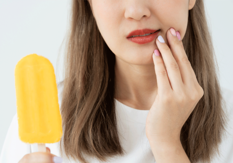 A close-up of a person holding a yellow popsicle in their left hand while their right hand touches their lips. The person has shoulder-length brown hair and is wearing a white shirt.