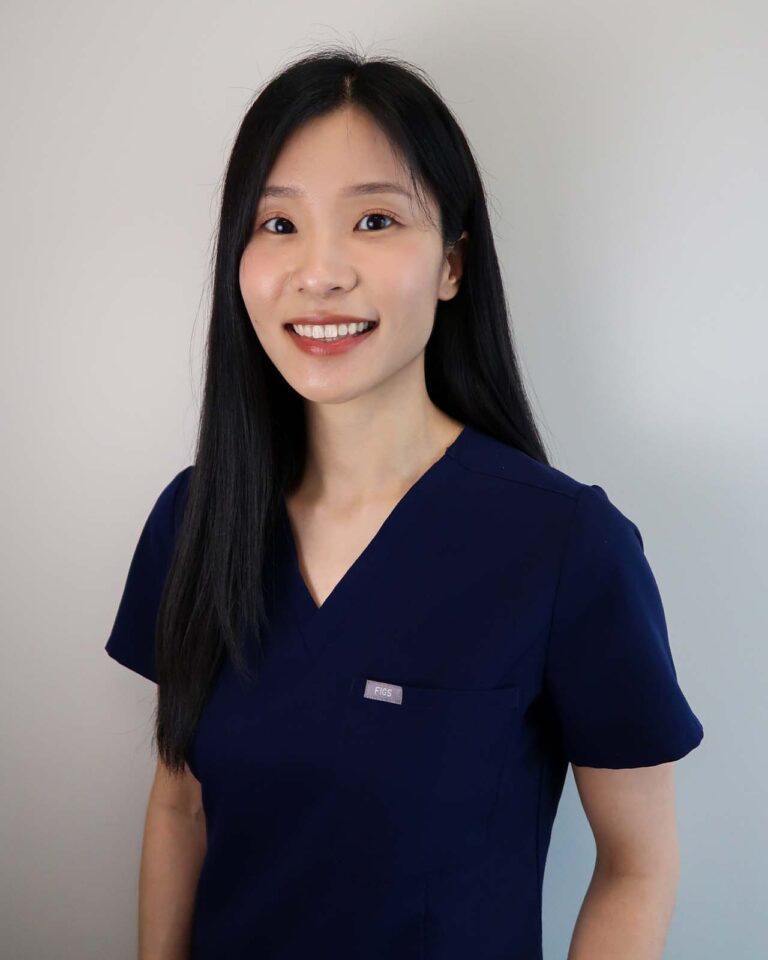 A woman wearing a dark blue scrub top associated with medical professionals, standing against a plain light-colored background.
