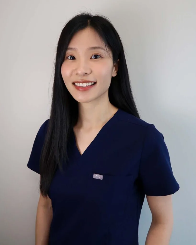 A woman wearing a dark blue scrub top associated with medical professionals, standing against a plain light-colored background.