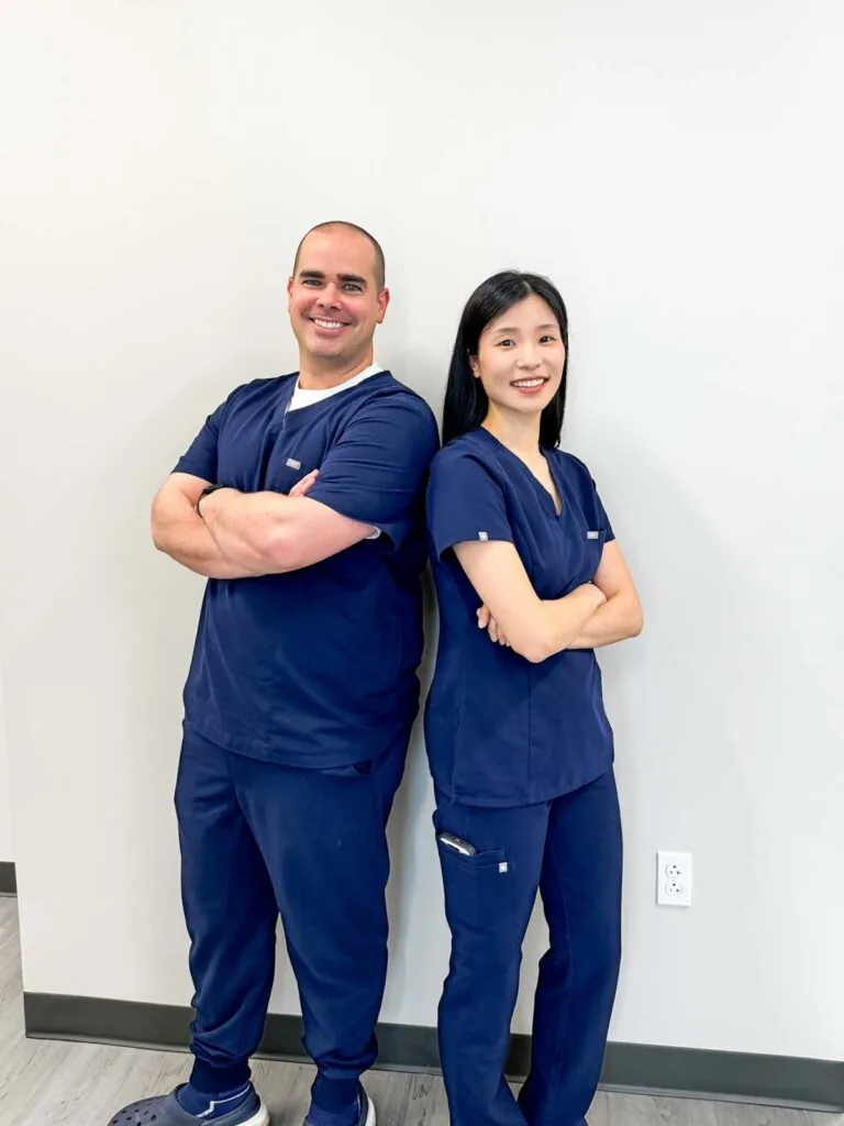 Two individuals in dark blue medical scrubs standing side by side with arms crossed. The background is a plain light-colored wall with a white electrical outlet near the bottom right corner.