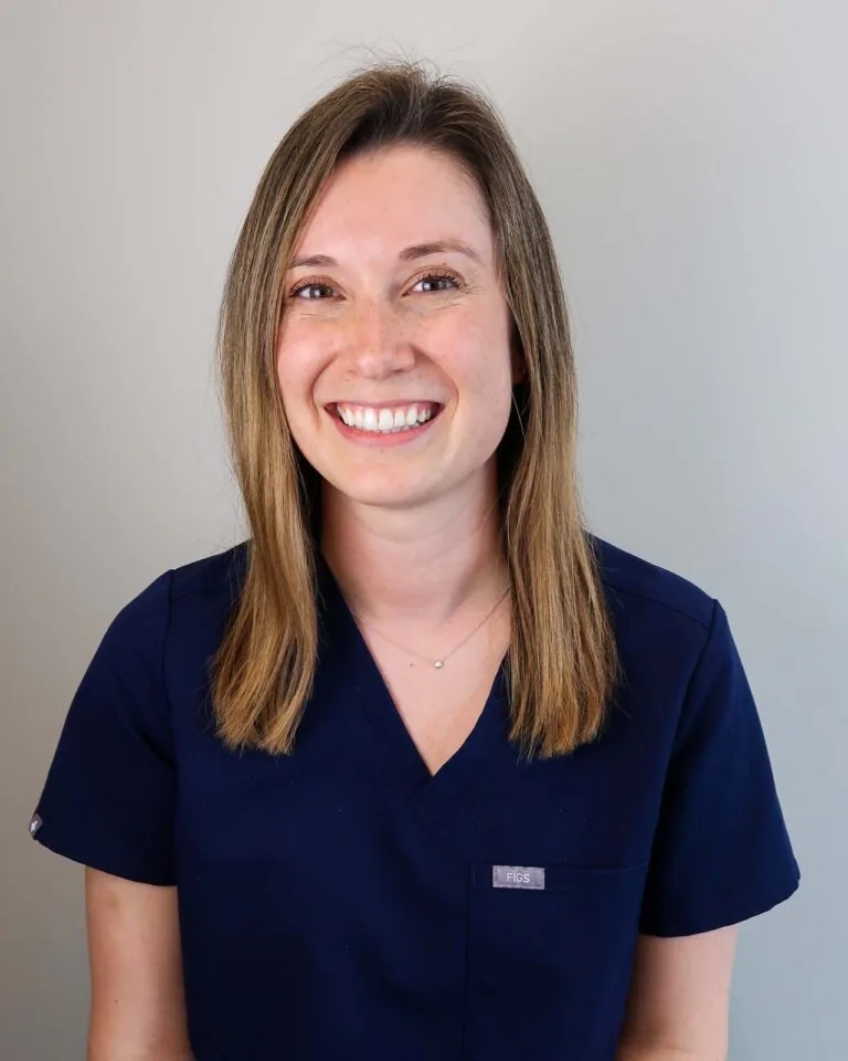 A woman wearing a dark blue scrub top associated with medical professionals, standing against a plain light-colored background.