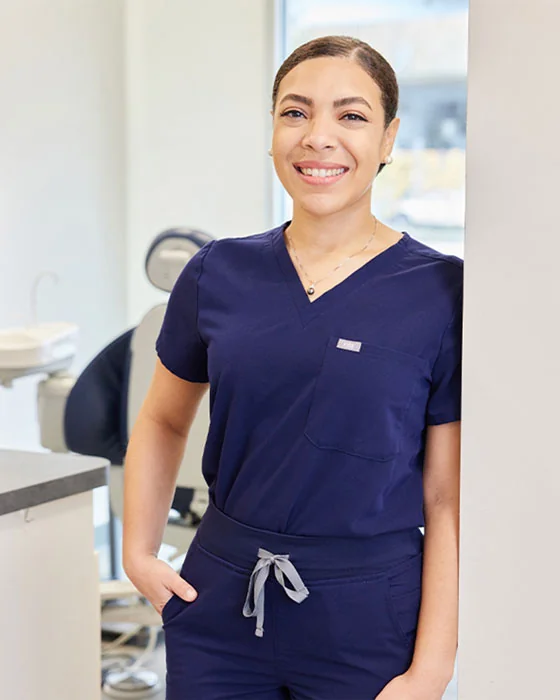 A woman wearing navy blue medical scrubs, standing in a clinical setting with medical equipment and a chair in the background.